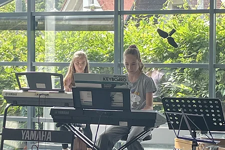 Zwei Mädchen spielen Keyboard auf einer Bühne vor großen Fenstern mit Blick auf grüne Pflanzen und Häuser.