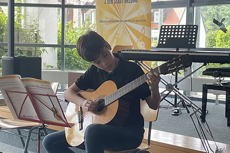 Junge sitzt auf Stuhl und spielt Gitarre vor Notenständer in Musikschule Greding.