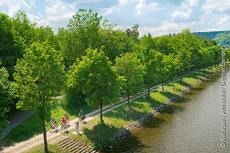 Fünf Radfahrer auf einem Weg entlang eines Flusses mit Bäumen und Hügeln im Hintergrund bei bewölktem Himmel.