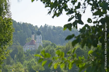 Ein Pärchen wandert unterhalb der Wallfahrtskirche Breitenbrunn entlang satter grüner Wiesen. Die Wallfahrtskirche ist von grünen Wäldern umgeben.