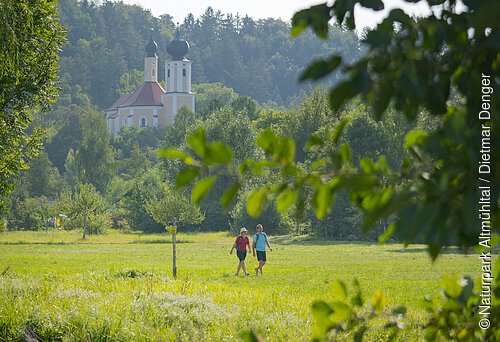 Ein Pärchen wandert unterhalb der Wallfahrtskirche Breitenbrunn entlang satter grüner Wiesen. Die Wallfahrtskirche ist von grünen Wäldern umgeben.