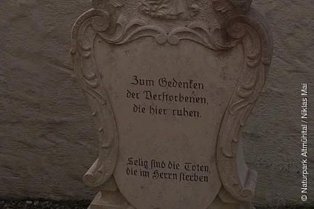 Grabstein mit Relief einer stehenden Figur und der Inschrift „Zum Gedenken der Verstorbenen, die hier ruhen“ in deutscher Sprache.