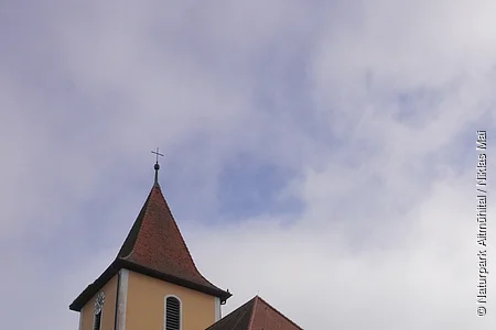 Kirche mit gelber Fassade, rotem Dach und Turm mit Kreuz vor bewölktem Himmel.