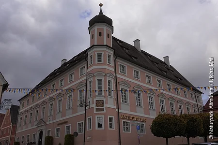 Eckgebäude mit Turm und Schriftzug „Antiquitäten“ unter bewölktem Himmel, bunte Wimpelkette gespannt.