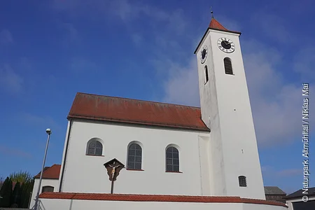 Kirchturm mit Uhr und weißes Kirchengebäude unter blauem Himmel, Vorderseite mit drei Fenstern und Kreuz am Gebäude.