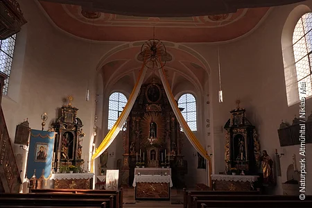 Innenraum einer Kirche mit Altar, zwei Fenstern und hängenden gelben Stoffbahnen am Altarbereich.