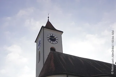 Kirchturm mit Uhr und weißem Gebäude bei bewölktem Himmel in einem Dorf.
