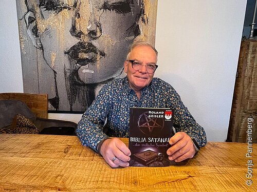 Mann mit Brille sitzt an Holztisch und hält Buch „Biblia Satanae“ von Roland Geisler hoch.