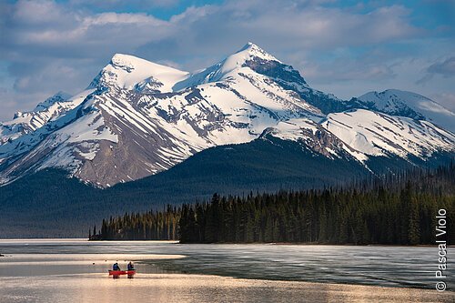 Zwei Personen in einem roten Kanu auf einem See vor schneebedeckten Bergen und einem Nadelwald.