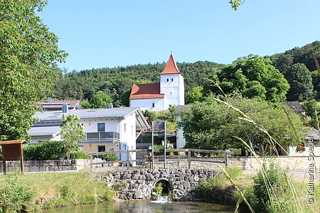 Fluss mit klarstem Wasser, Brücke aus Stein, Häuser und Kirche mit rotem Dach vor bewaldetem Hügel unter blauem Himmel.
