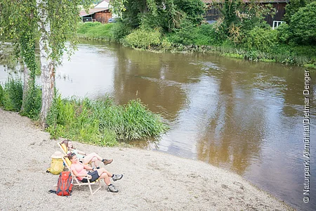 Zwei Personen mit Rucksäcken neben sich  sitzen auf Liegestühlen am Flussufer neben Bäumen an einem sonnigen Tag.