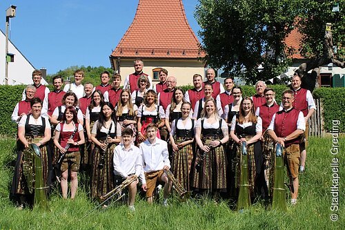 Stadtkapelle Greding Gruppenfoto einer Blaskapelle mit 31 Personen in Trachten vor Haus mit rotem Dach bei Sonnenschein auf Wiese.