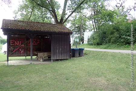 Holzunterstand mit Bank und Graffiti, daneben Mülltonnen und Waldweg im Grünen