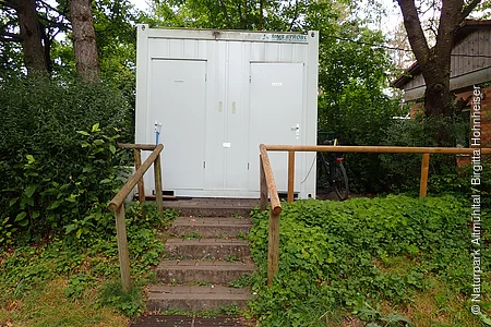 Außenansicht eines weißen Containers mit zwei Türen, beschriftet mit „Herren“ und „Damen“, umgeben von Grün und Bäumen.
