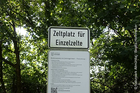Schild mit der Aufschrift „Zeltplatz für Einzelzelte“ und weiteren Informationen vor grünem Laub.
