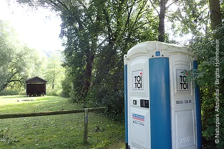 Mobiles Toilettenhäuschen mit blau-weißer Außenwand steht am Waldrand auf einer Wiese neben einem Holzzaun.