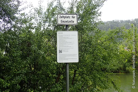 Schild mit der Aufschrift „Zeltplatz für Einzelzelte“ vor Büschen und Flussufer im Hintergrund.