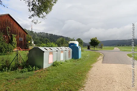 Recyclingcontainer neben einem Feldweg, links ein Holzanbau und im Hintergrund Hügel und bewölkter Himmel.