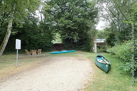 Grüner Kanu und Kajak auf Grasfläche neben Waldweg mit Sitzgruppe und Brücke im Hintergrund.