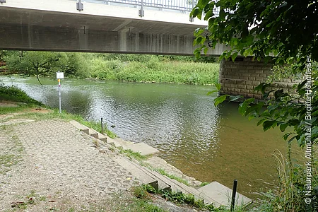 Uferbereich mit Stufen am Fluss unter einer Betonbrücke, umgeben von Grünpflanzen und Bäumen.