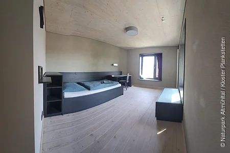Schlicht eingerichtetes Zimmer mit Einzelbett, Schreibtisch, Stuhl, Fenster und Holzboden.