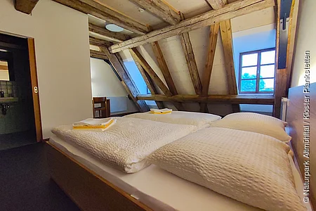 Schlafzimmer mit Dachschräge, Holzbalken, zwei Betten mit weißen Decken und Kissen, Fenster und offenem Badezimmer.