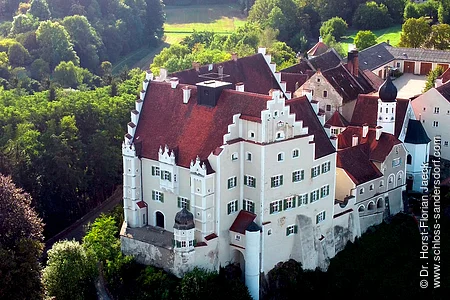 Luftaufnahme eines historischen Schlosses mit roten Dächern, weißen Mauern und umliegendem Wald.