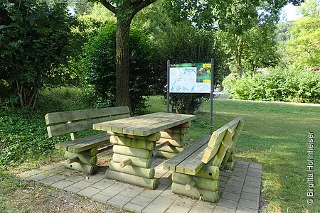 Holztisch mit zwei Bänken auf gepflastertem Platz unter Baum, im Hintergrund Infotafel und Grünfläche.