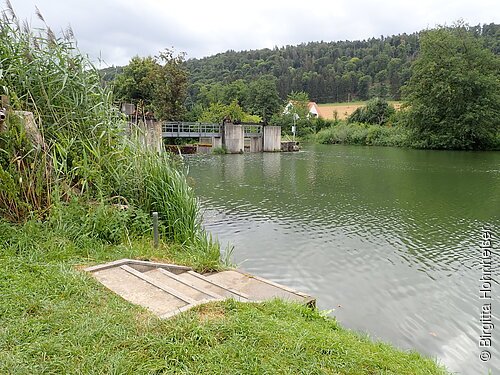 Kottingwörther Wehr Ufer mit Treppe am Fluss, im Hintergrund Schleusenanlage und bewaldete Hügel unter bewölktem Himmel.
