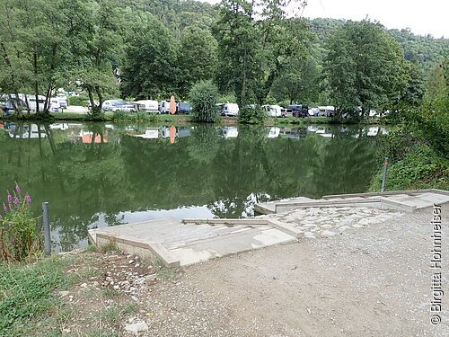 Anlegestelle Kratzmühle Ufer mit Treppe an einem ruhigen See, gegenüber Bäume und Wohnwagen auf einer Wiese.
