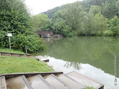 Treppe führt zu einem ruhigen Fluss mit grünem Ufer und Bäumen im Hintergrund bei bewölktem Himmel.