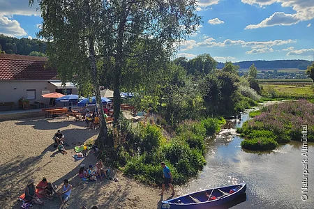 Menschen sitzen am Ufer eines Flusses mit Kanus, Bäumen und einem Gebäude mit Sonnenschirmen im Hintergrund.