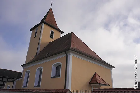 Kirchturm mit spitzem Dach und gelber Fassade unter bewölktem Himmel, von unten fotografiert.