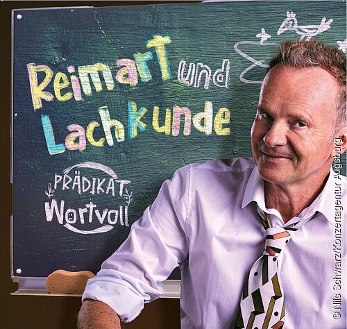 Willy Astor "Reimart und Lachkunde" Willy Astor sitzt in einem fliederfarbenem Hemd mit gemusterter Krawatte vor einer Schultafel mit der Aufschrift: Reimart und Lachkunde - Prädikat Wortvoll