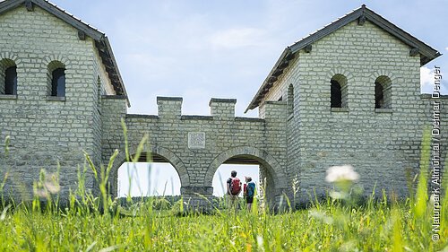 Römerkastell Vetoniana bei Pfünz Zwei Personen mit Rucksäcken stehen unter einem steinernen Torbogen einer historischen Anlage bei sonnigem Himmel.