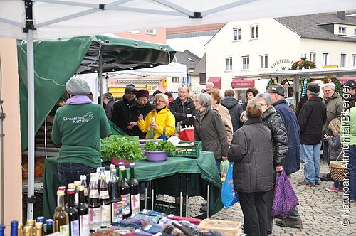Denkendorfer Dorfmarkt Gemüsestand mit Verkäuferin im grünen Pullover von hinten. Daneben stehen verschiedene Obstsaftflaschen. Der Stand steht auf dem Dorfplatz, umgeben von einer größeren Personengruppe. Im Hintergrund sieht man noch andere Marktstände.